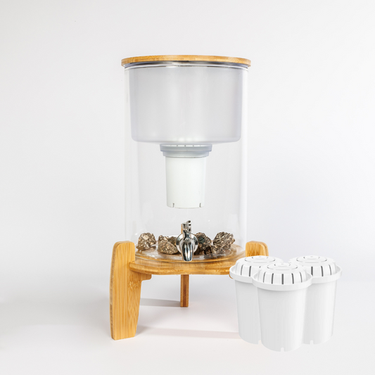 Glazen waterfilter met houten standaard en RVS kraan. Bovenin een glazen opvangtank met filterhouder. Onderaan mineralenstenen. Naast het filter staan drie witte reservefilter cartridges, samen voldoende voor een half jaar gebruik.