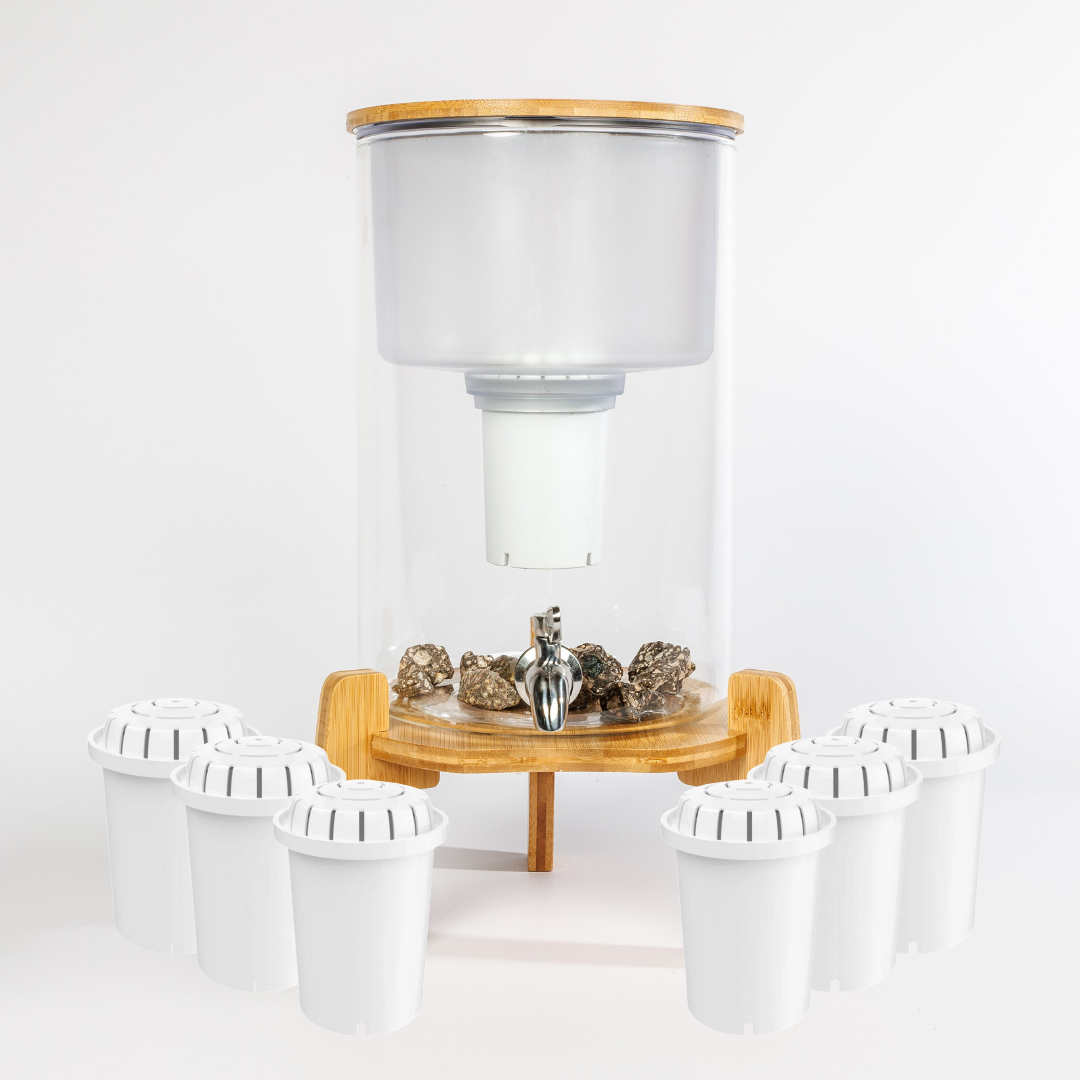 Glazen waterfilter met houten standaard en RVS kraan. Bovenin een glazen opvangtank met filterhouder. Onderaan mineralenstenen. Rondom het filter staan zes witte reservefilter cartridges, samen voldoende voor één jaar gebruik.